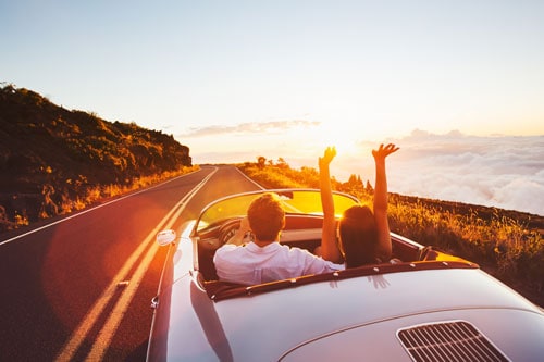 man and woman driving in convertible into the sunset | Labor Day Weekend Travel Safety Tips